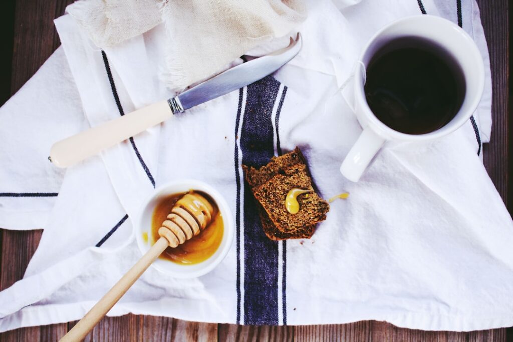 A jar of honey with a wooden honey dipper, a slice of dark bread, and a cup of coffee on a white linen cloth