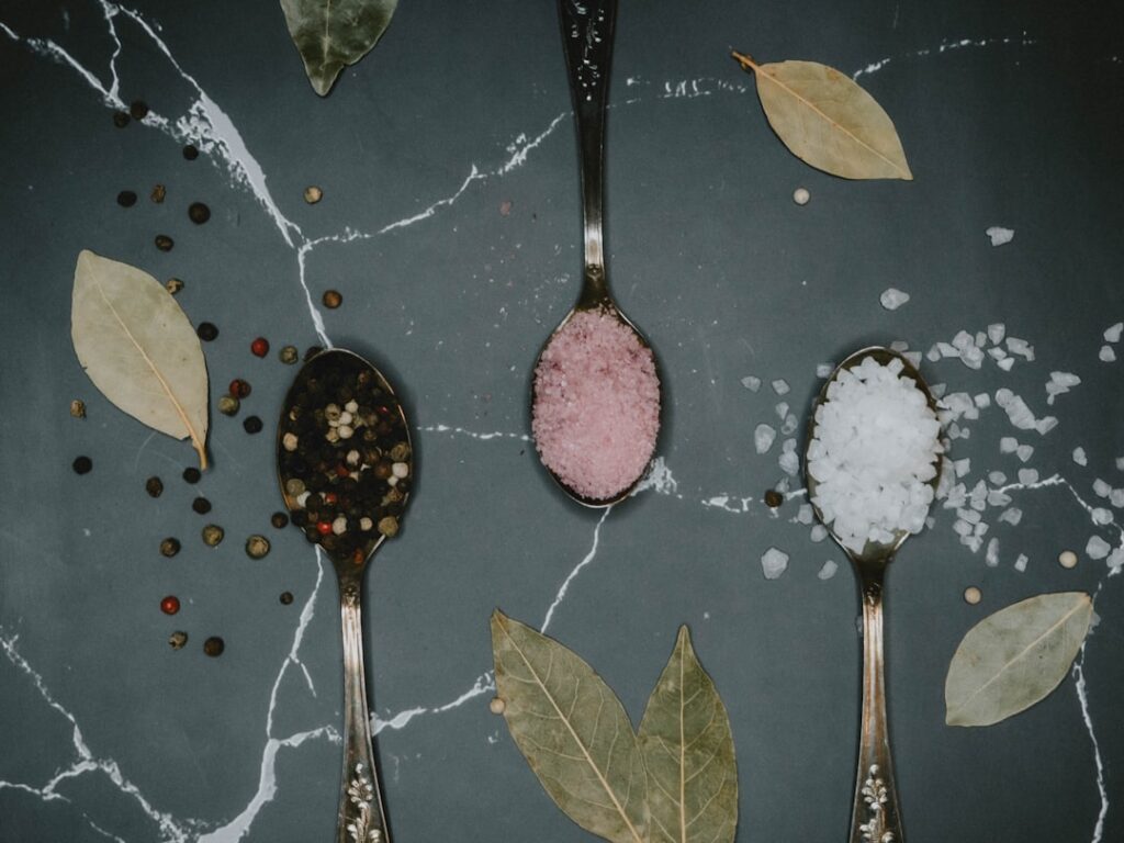 Three spoons holding different varieties of salt — black pepper blend, pink Himalayan, and coarse sea salt — on a marble surface with dried bay leaves