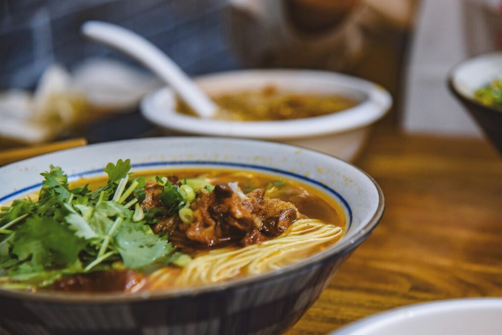 A steaming bowl of Chinese noodle soup with rich broth, meat, and herbs served with chopsticks