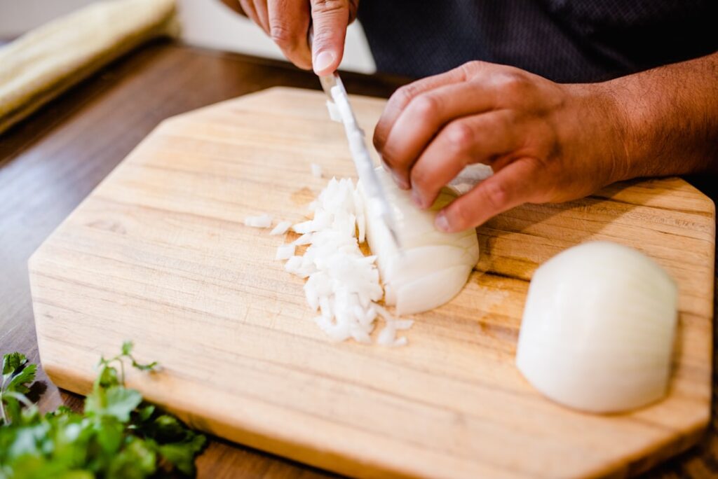 Close-up of hands dicing a white onion on a wooden cutting board, showing the moment cells are ruptured and the alliinase cascade begins