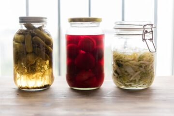 Three glass jars of fermented foods including pickles, pickled beets, and sauerkraut on a wooden surface